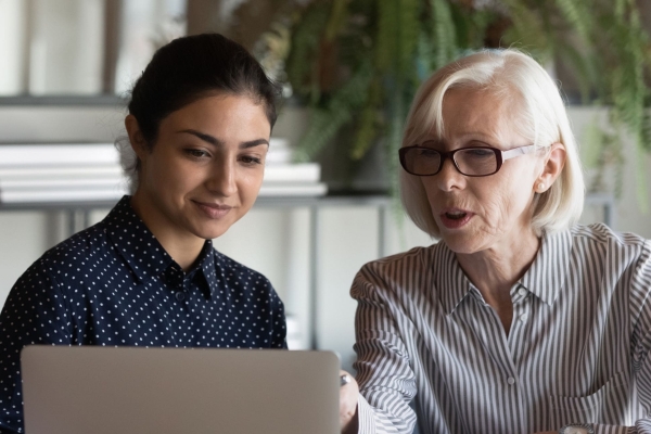 A young Asian woman with tied back dark hair and wearing a blue top, is sat looking at a laptop next to a white older woman with collar length white-bonde hair. She wears glasses and a light shirt. They are sat in an office environment in front of pl